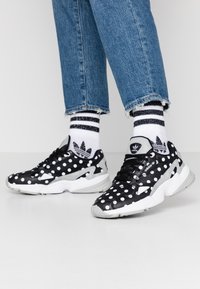 Svart och vit prickig sneakers med en texturerad yta, med grå accenter och en vit sula, tillsammans med randiga strumpor och jeans.