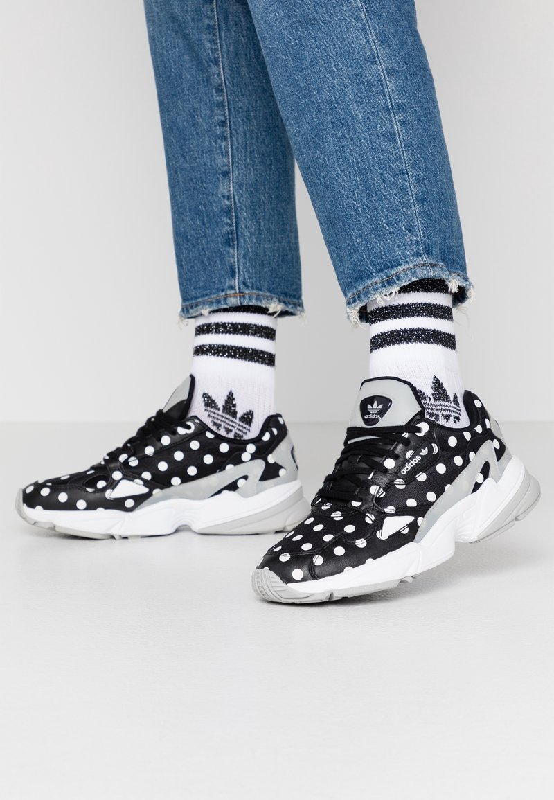 Svart och vit prickig sneakers med en texturerad yta, med grå accenter och en vit sula, tillsammans med randiga strumpor och jeans.