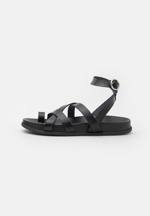 Las sandalias de piel negra cuentan con amplias correas entrelazadas y una correa en el tobillo con hebilla. Suela plana con una textura suave y diseño abierto.