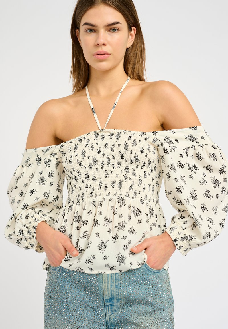 Top a spalla scoperta con motivo floreale, corpino elastico arricciato, maniche a sbuffo e spalline sottili in crema e nero. Indossato con pantaloncini di jeans.