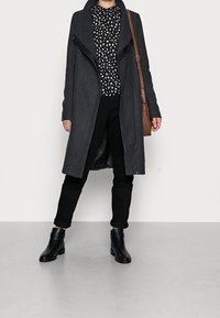 Cappotto in misto lana grigio con zip asimmetrica, indossato sopra una blusa bianco e nera a pois, abbinato a pantaloni neri e stivaletti.