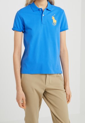 Personne portant un polo bleu vif à manches courtes avec un logo jaune de cheval et cavalier, associé à un pantalon beige, debout devant un fond uni.
