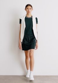 Top corto de color verde oscuro con ribete blanco, combinado con una falda a juego. Un chaleco blanco está caído sobre los hombros; estilizado con zapatillas blancas.