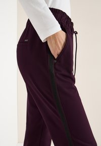 Fialové sportovní kalhoty s elastickým pasem, bočními kapsami a černým vertikálním pruhem. Hladká textura s upnutým designem.