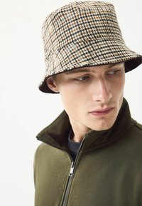 Brown and beige houndstooth bucket hat made of woven fabric; features a wide brim and a black interior band. Model wears an olive jacket.