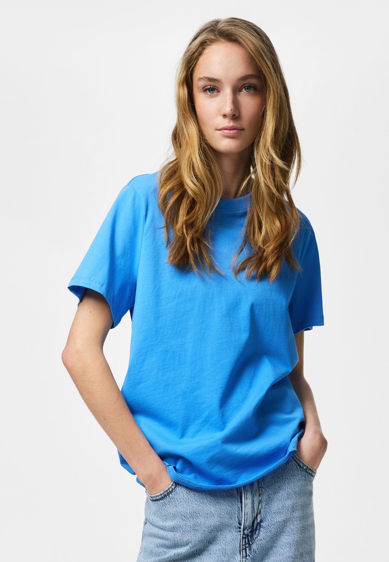 Jonge vrouw met lang blond haar, die een felblauw T-shirt en lichtblauwe spijkerbroek draagt, met handen in de zakken, tegen een effen witte achtergrond.