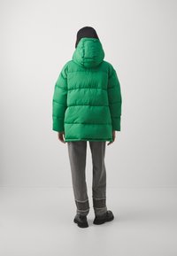 Grüne Puffjacke mit Kapuze, die ein gestepptes Design aufweist. Der Rücken hat eine lockere Passform und dicke Polsterung. Getragen mit grauen, leicht abgetragenen Hosen.