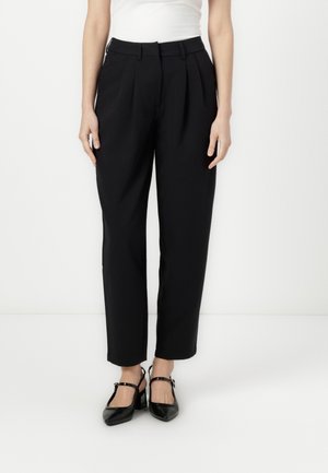 Kvinde iført sorte, højtaljede, plisserede bukser og sorte Mary Jane-hæle, stående mod en hvid baggrund.