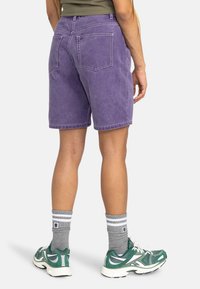 Shorts in denim viola con una vestibilità comoda, cinque tasche e dettagli di cucitura visibili. Indossati con calzini a righe grigi e sneaker verdi.