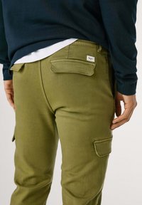 Olivenfarvede cargo bukser med slim fit, der har baglommer og et lille brandmærke. Stoffet har en glat, mat tekstur.