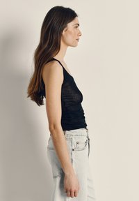 Žena s dlhými hnedými vlasmi, oblečená v čiernom tielku bez rukávov a svetlomodrých džínsoch, stojaca v profile na jednoduchom pozadí.