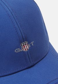 Modrá bejzbalová čiapka vyrobená z textilného materiálu. Obsahuje vyšívané logo so štítovým designom v červenej, bielej a modrej farbe.