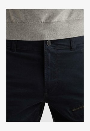 Povečan posnetek temno modrih hlač z gumbno zaponko, zankami za pas, z žepom z zadrgo na desnem stegnenem delu in pasom bež puloverja, zataknjenim v hlače.