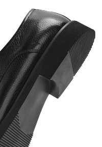 Zbliżenie obcasa czarnego skórzanego buta wizytowego z fakturowaną gumową podeszwą i średniej wysokości blokowym obcasem na białym tle.