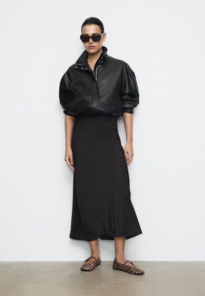 Veste en cuir noir à manches bouffantes, jupe midi noire et ballerines à imprimé léopard. La mannequin porte des lunettes de soleil sombres. Textures lisses et design épuré.