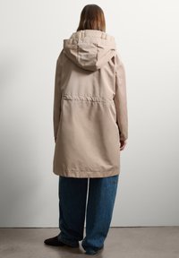 Parka beige impermeabile con cappuccio, foderato in tessuto trapuntato e vita cintrata. Indossato sopra jeans a gamba larga blu, visto da dietro.