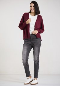 Bordeauxfarbener Strickcardigan über einem weißen Oberteil, kombiniert mit grauen Jeans. Das Modell trägt weiße Sneakers mit braunen Akzenten. Einfaches Design, lässige Passform.