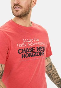 Rød bomulds t-shirt med korte ærmer og rund hals, med trykt tekst der står "Lavet Til Daglige Eventyr" og "FORFØLG NYE HORISONTER."