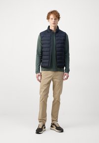Marineblaue Steppweste über einem dunkelgrünen Langarmshirt, kombiniert mit beigen Hosen und schwarzen Sneakers mit braunen Akzenten. Glatte Texturen.