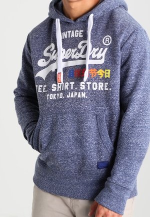 Homme portant un sweat à capuche SuperDry bleu avec cordons blancs et texte imprimé Tokyo, Japon, mains dans les poches, sur un fond uni.