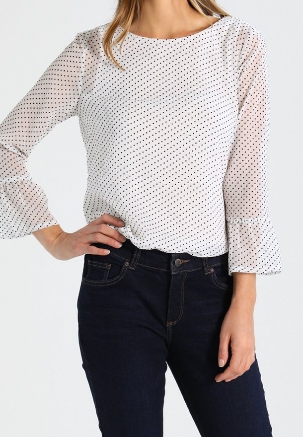 Blusa bianca con pois neri, maniche lunghe con polsini a campana, abbinata a jeans scuri. La blusa presenta un colletto tondo e un materiale trasparente.