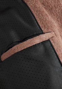 El tejido de fieltro marrón texturizado contrasta con el material negro liso que presenta un patrón perforado; se puede ver un bolsillo integrado en el lado negro.
