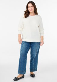 Maglione in tessuto a crema, vestibilità morbida; jeans a gamba larga blu con cuciture a contrasto; ballerine nere con dettagli decorativi.