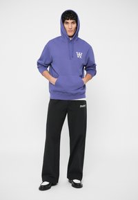 Fialová mikina s predným vreckom a logom, v kombinácii s čiernymi nohavicami širokého strihu s brandingom "Doubler". Model má na sebe topánky s kontrastnými akcentmi.