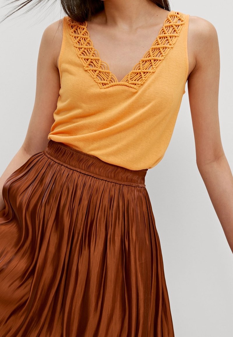 Top arancione senza maniche con rifiniture in pizzo decorativo su un collo a V, abbinato a una gonna plissettata marrone, con un elastico arricciato in vita.