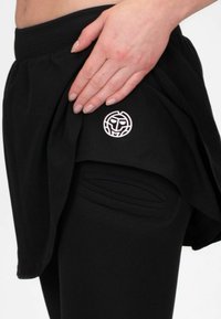Pantalones cortos atléticos negros con una cintura alta, que presentan un parche de logo y un diseño en capas. Textura suave, corte holgado y bolsillos laterales.