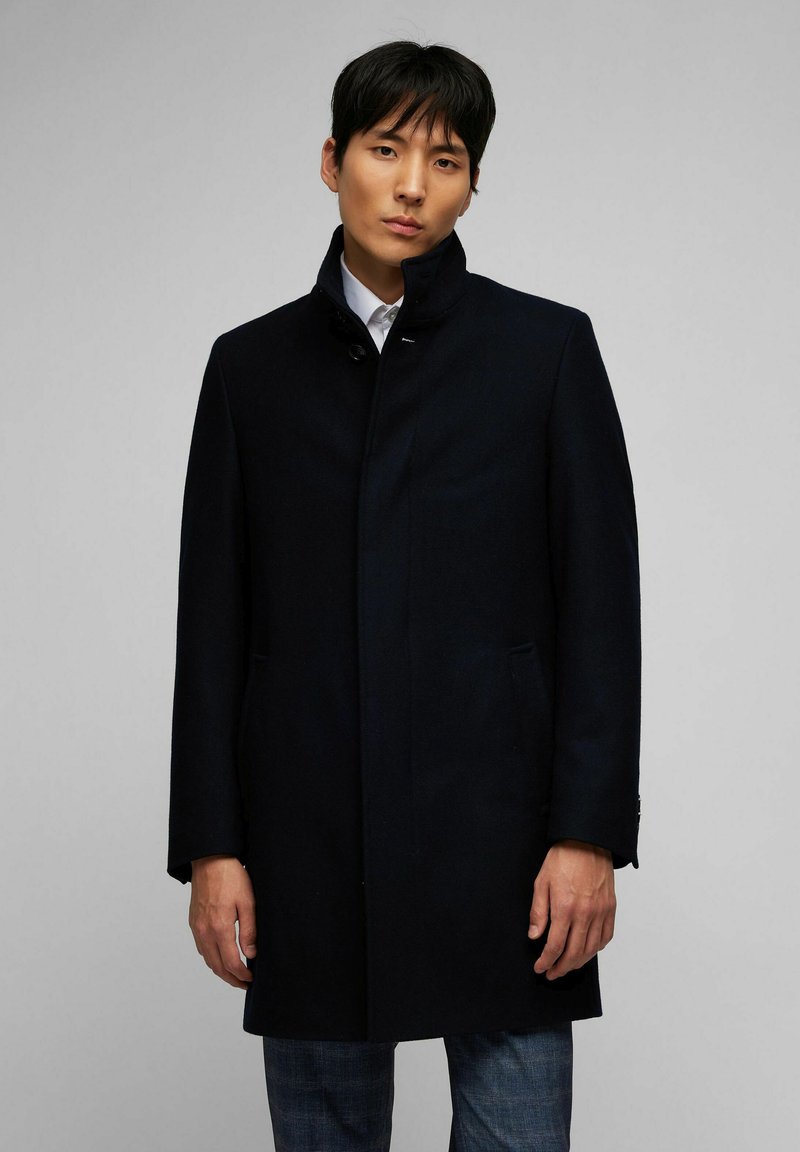 Cappotto di lana blu navy con colletto alto, chiusura frontale a bottoni e tasche laterali, abbinato a una camicia bianca e jeans scuri.