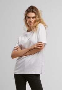 Tricou alb din bumbac, cu o croială lejeră, având un mic logo negru pe piept. Mâneci scurte și decolteu rotund. Fără modele sau accente.