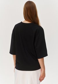 Czarna, oversize t-shirta z krótkim rękawem, wykonana z miękkiego materiału. Posiada okrągły dekolt i luźny krój, noszona na białej spódnicy.