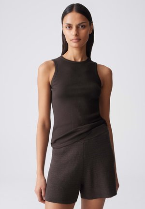Young woman with straight dark hair wearing a sleeveless dark ribbed top and textured dark shorts, standing against a plain background.