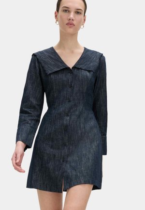 Vrouw die een donker denim jurk met knopen draagt, met lange mouwen en een brede kraag, staand tegen een effen lichte achtergrond.