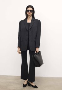 Blazer nero oversize con pantaloni slim abbinati. Occhiali da sole neri spessi, scarpe nere basse e una borsa a tracolla nera completano il look. Tessuto dalla texture liscia.