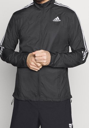Mężczyzna w czarnej kurtce Adidas na zamek z białymi paskami na rękawach oraz czarnych spodenkach Adidas, stoi ze splecionymi rękami.