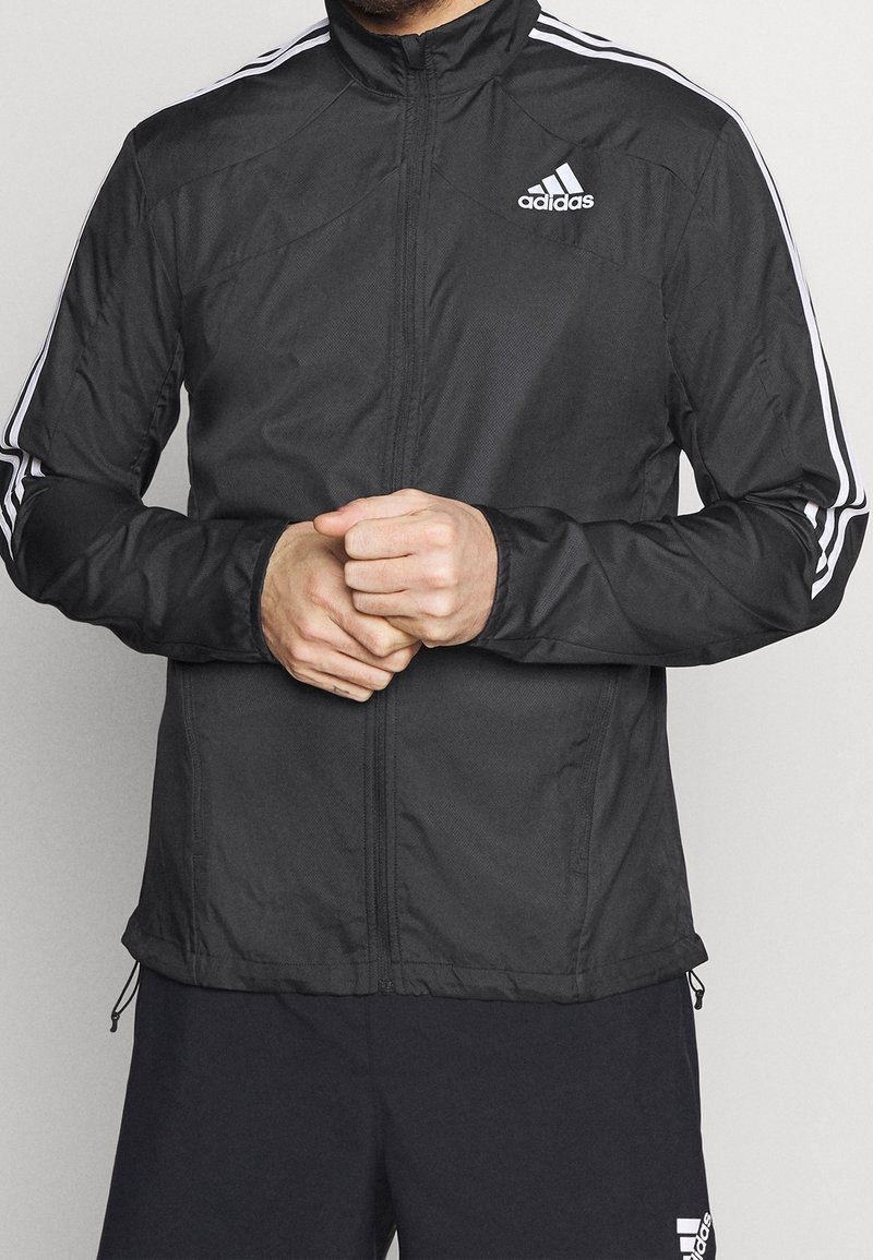 Homme portant une veste zippée noire Adidas avec des bandes blanches sur les manches et un short noir Adidas, debout avec les mains jointes.