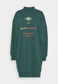 Zelené šaty s dlouhými rukávy a vysokým límcem, se žebrovanými manžetami. Obsahují text a logo v žluté a červené barvě. Měkký materiál.