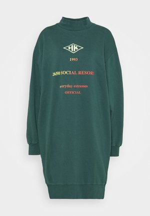 Zelené šaty s dlouhými rukávy a vysokým límcem, se žebrovanými manžetami. Obsahují text a logo v žluté a červené barvě. Měkký materiál.