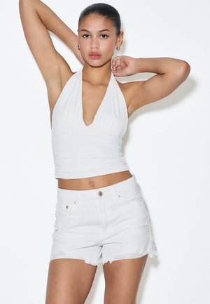 Žena so zviazanými vlasmi, oblečená v bielom halter top a bielych rifľových šortkách, pózuje s rukami zdvihnutými na bielom pozadí.