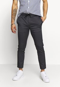 Hombre con pantalones de rayas marino y cintura con cordón, zapatillas blancas y una camisa de rayas azul y blanca de manga corta, de pie con las manos en los bolsillos.