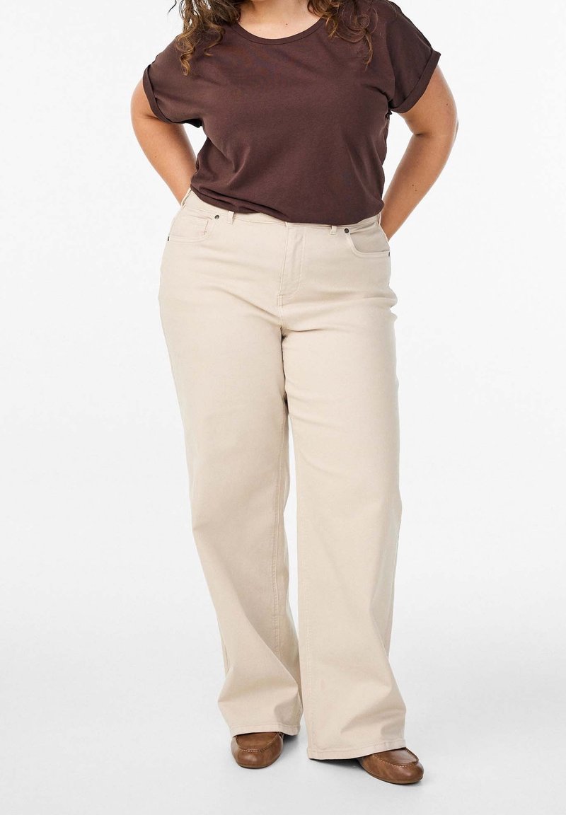 Femme portant une chemise marron à manches courtes, un pantalon beige large et des chaussures marron, debout les mains dans le dos sur fond blanc.