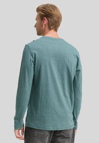 Camisa de manga larga color verde azulado con un escote redondo y una textura suave, que presenta un sutil detalle de costura en los hombros y los lados.
