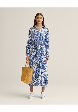 Kvinde iført en hvid og blå blomsterprintet lang kjole med bånd i taljen, hvide sneakers og bærende en stor tanfarvet vævet taske.