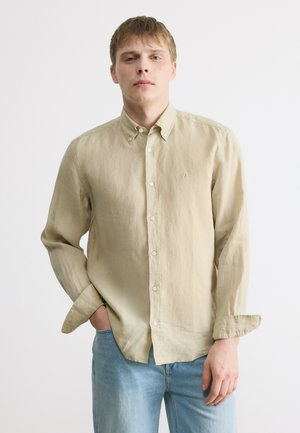 Młody mężczyzna w beżowej koszuli z kołnierzykiem zapinanej na guziki i niebieskich dżinsach, stojący z jedną ręką w kieszeni na białym tle.