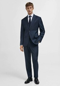 Completo blu navy realizzato in tessuto strutturato, con giacca a un bottone, pantaloni a gamba dritta e cravatta abbinata. Scarpe nere lucide.
