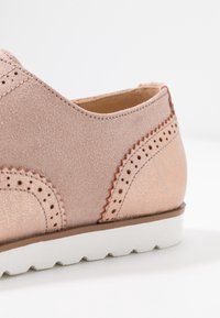 Vaaleanpunainen mokkanahkainen kenkä, jossa koristeelliset rei'itykset ja metallinen yksityiskohta. Valkoinen pohja, jossa on teksturoidut reunat.