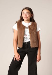 Gilet en fausse fourrure marron avec une texture douce, à l'avant ouvert, porté par-dessus un t-shirt graphique blanc, associé à un pantalon large noir.