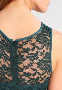 Dunkelblaues Kleid mit Spitzenrücken, Blumendesign und Reißverschluss. Es hat einen gewellten Ausschnitt und glattes Material an den Schultern.